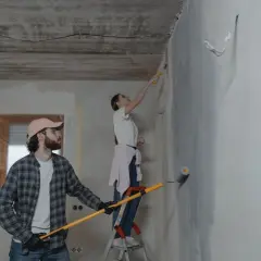 Para maluje ściany wałkiem, podczas gdy mężczyzna w czapce przygotowuje się do malowania pistoletem.