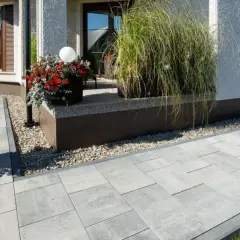 Nowy, nowoczesny chodnik z szarej kostki brukowej prowadzi do domu. Wokół rosną rośliny w donicach.