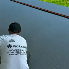 Pracownik w koszulce z napisem "Posadzki żywiczne" wykonuje prace na ciemnej, gładkiej powierzchni.