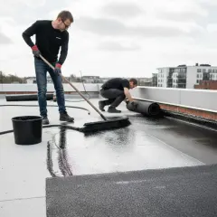 Dwóch mężczyzn układa abizol na dachu. Jeden rozwałkowuje materiał, drugi zamiata.