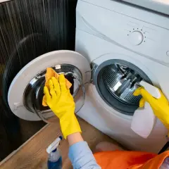 Ręce w żółtych rękawiczkach czyszczą szklane drzwiczki pralki. Dowiedz się, czym wyczyścić pralkę, aby była jak nowa.