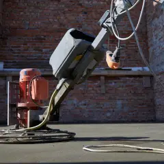 Robot do zacierania betonu pracuje na budowie. Robotnik w niebieskich spodniach kieruje maszyną, która wygładza świeżą posadzkę.