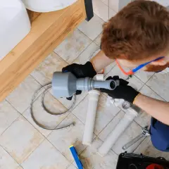 Instalator pracuje nad odpowietrzeniem kanalizacji, montując elementy syfonu pod umywalką.