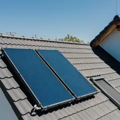 Dwa panele **solary** na szarym dachu pod błękitnym niebem. Obok okno dachowe.