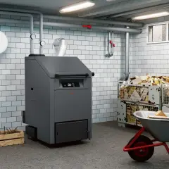 Kotłownia z nowoczesnym piecem na biopaliwo, obok składowane drewno opałowe w metalowych koszach i taczka z polanami.