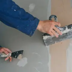 Ręce w niebieskim kombinezonie nakładają masę gipsową na ścianę za pomocą szpachli.