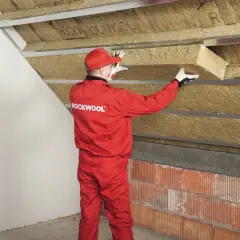Pracownik w czerwonym kombinezonie Rockwool montuje izolację na ścianie szczytowej poddasza.
