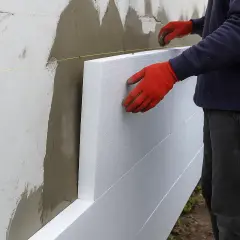 Pracownik w czerwonych rękawicach montuje płyty styropianowe na ścianie domu, tworząc izolację elewacji.