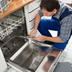 Fachowiec w niebieskim kombinezonie dokonuje podłączenia zmywarki, pracując przy jej wnętrzu. Obok leżą narzędzia.