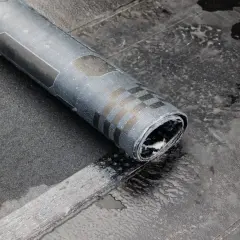 Zrolowana papa na zimno, z charakterystycznym wzorem szachownicy, leży na mokrej, ciemnej powierzchni.
