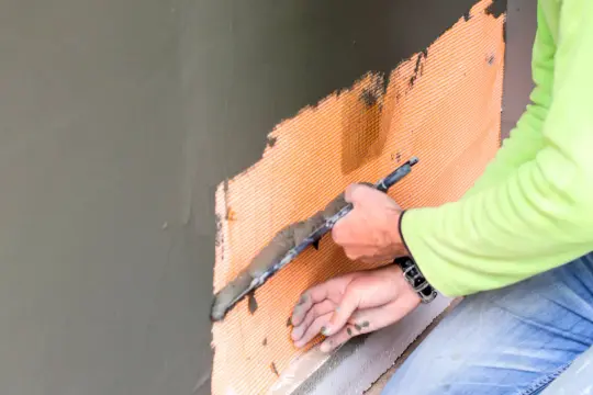 Ręce pracownika w zielonej bluzie i dżinsach wykonują tynkowanie na pomarańczowej siatce zbrojeniowej, używając kielni.