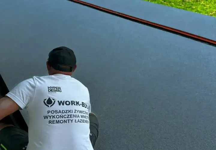 Pracownik w koszulce z napisem "Posadzki żywiczne" wykonuje prace na ciemnej, gładkiej powierzchni.