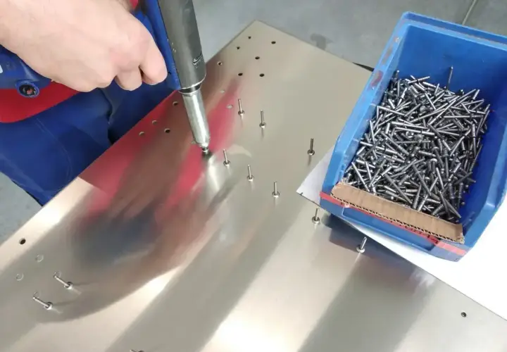 Ręka pracownika używa narzędzia do montażu nitów zrywalnych na metalowej powierzchni. Obok znajduje się niebieskie pudełko pełne nitów.