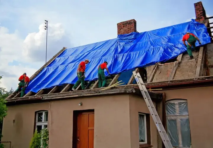 Pracownicy zabezpieczają dach niebieską plandeką. Remont dachu wymaga pozwolenia na budowę? Sprawdź przepisy.