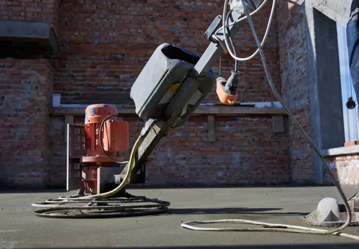 Robot do zacierania betonu pracuje na budowie. Robotnik w niebieskich spodniach kieruje maszyną, która wygładza świeżą posadzkę.
