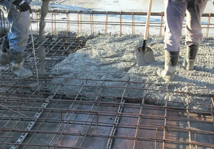 Betonowanie stropu. Robotnicy w gumowcach wylewają beton na zbrojenie. Łopata czeka na pomoc.