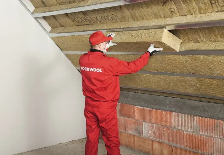 Pracownik w czerwonym kombinezonie Rockwool montuje izolację na ścianie szczytowej poddasza.