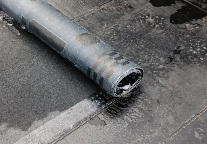 Zrolowana papa na zimno, z charakterystycznym wzorem szachownicy, leży na mokrej, ciemnej powierzchni.