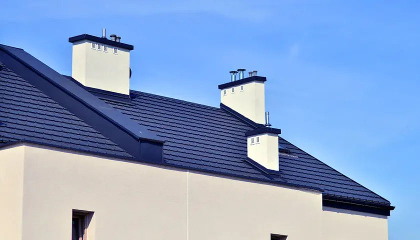 Nowy dom z ciemnym, dachówkowym dachem bezokapowym i białymi kominami pod błękitnym niebem.