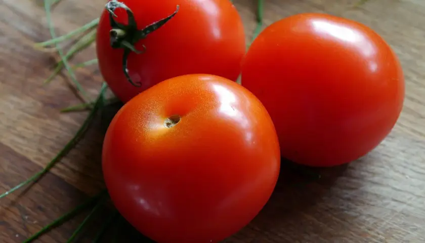 Trzy soczyste pomidory krakowskie na drewnianej desce, otoczone zielonymi szczypiorkami.