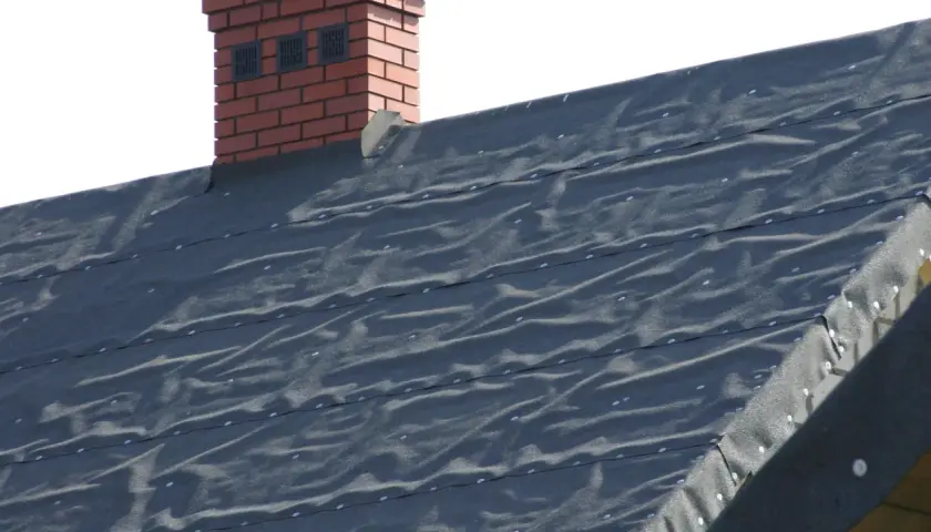 Ciemna papa bitumiczna na dachu, z widocznym fragmentem ceglanego komina.