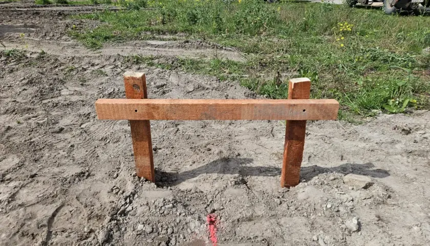 Drewniany palik z poprzeczką wyznacza przyszłą powierzchnię zabudowy. W tle widać plac budowy z maszynami.