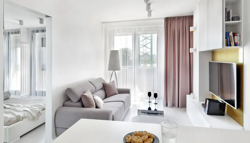 Jasny, bardzo mały salon w bloku z szarą kanapą, stolikiem kawowym i widokiem na balkon. Obok widać łóżko.