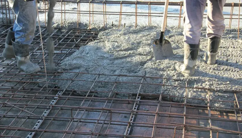 Betonowanie stropu. Robotnicy w gumowcach wylewają beton na zbrojenie. Łopata czeka na pomoc.