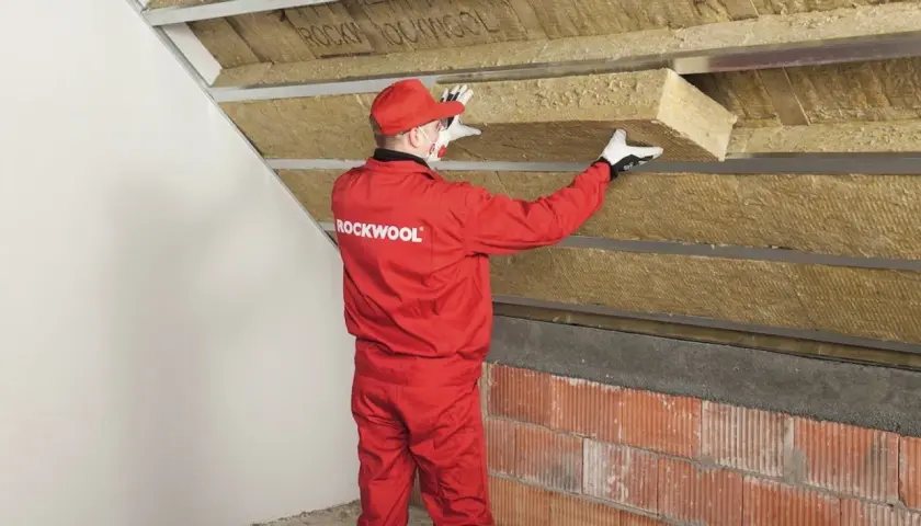 Pracownik w czerwonym kombinezonie Rockwool montuje izolację na ścianie szczytowej poddasza.