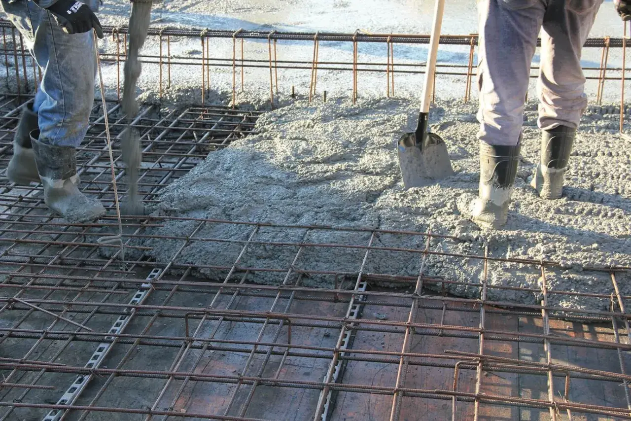 Betonowanie stropu. Robotnicy w gumowcach wylewają beton na zbrojenie. Łopata czeka na pomoc.