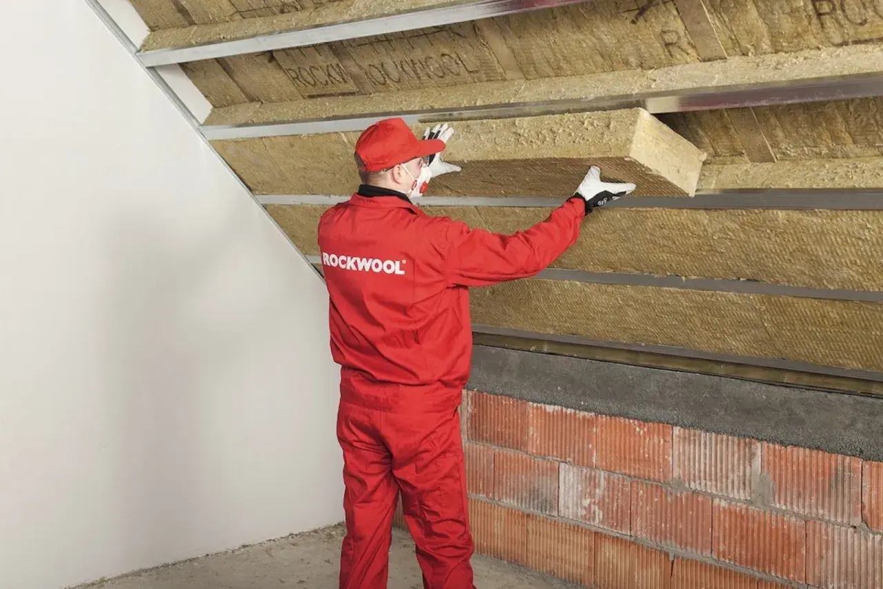 Pracownik w czerwonym kombinezonie Rockwool montuje izolację na ścianie szczytowej poddasza.