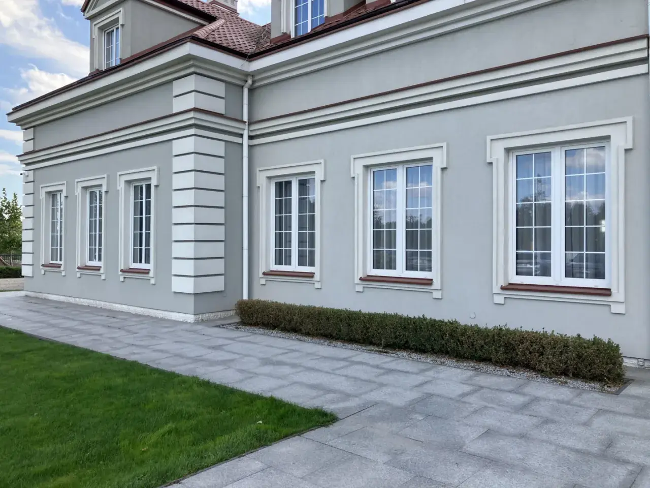 Elegancki budynek z jasnoszarym tynkiem, ozdobiony białymi pilastrami i gzymsami. Okna z białymi ramami i brązowymi parapetami.