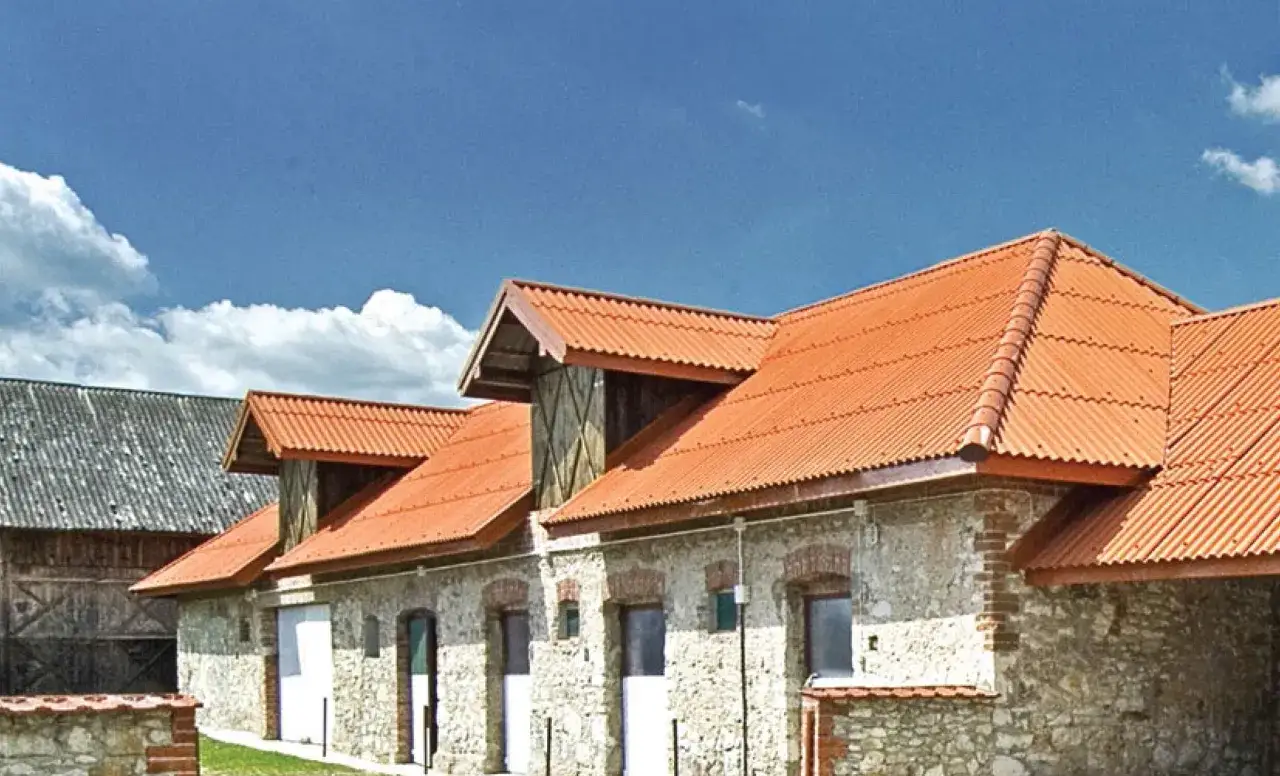 Stary budynek z szarym dachem, który może przypominać, jak wygląda azbest, obok nowszego z pomarańczową, falistą blachą.