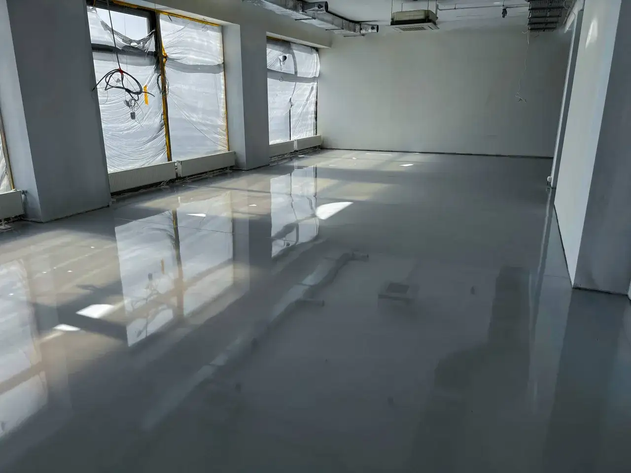 Lśniąca podłoga z żywicy w pomieszczeniu w budowie, odbijająca światło. Widać okna zasłonięte folią i instalacje na suficie.