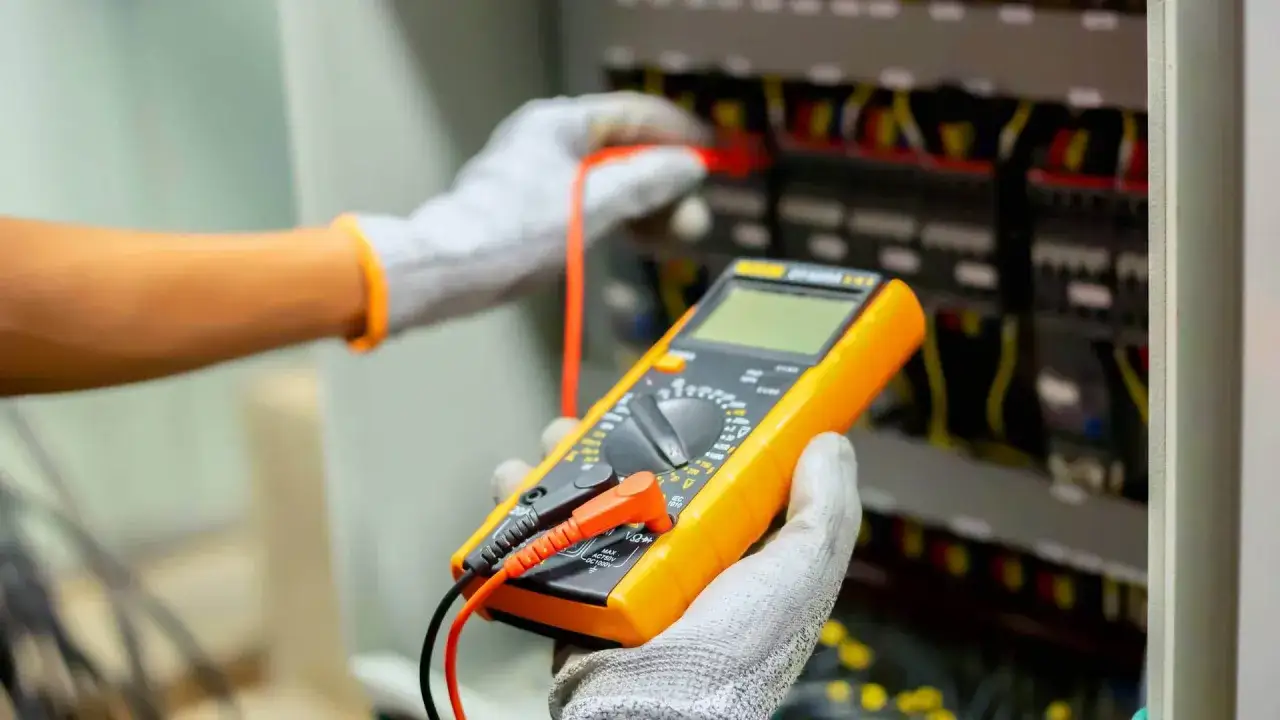Elektryk w rękawicach wykonuje przegląd instalacji elektrycznej za pomocą multimetru.