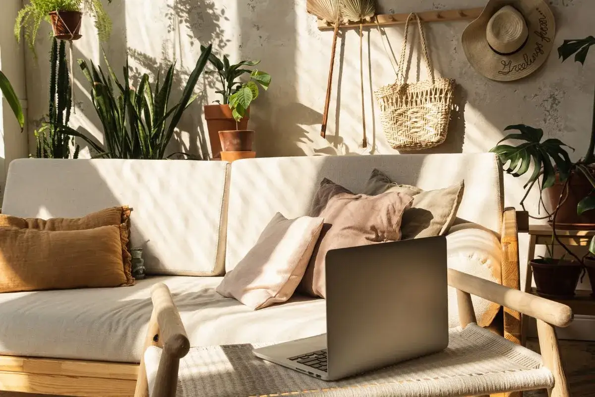 Słoneczny kącik z laptopem i mnóstwem roślin. Wygodna sofa, pleciona torba i kapelusz tworzą przytulny styl boho wnętrza.