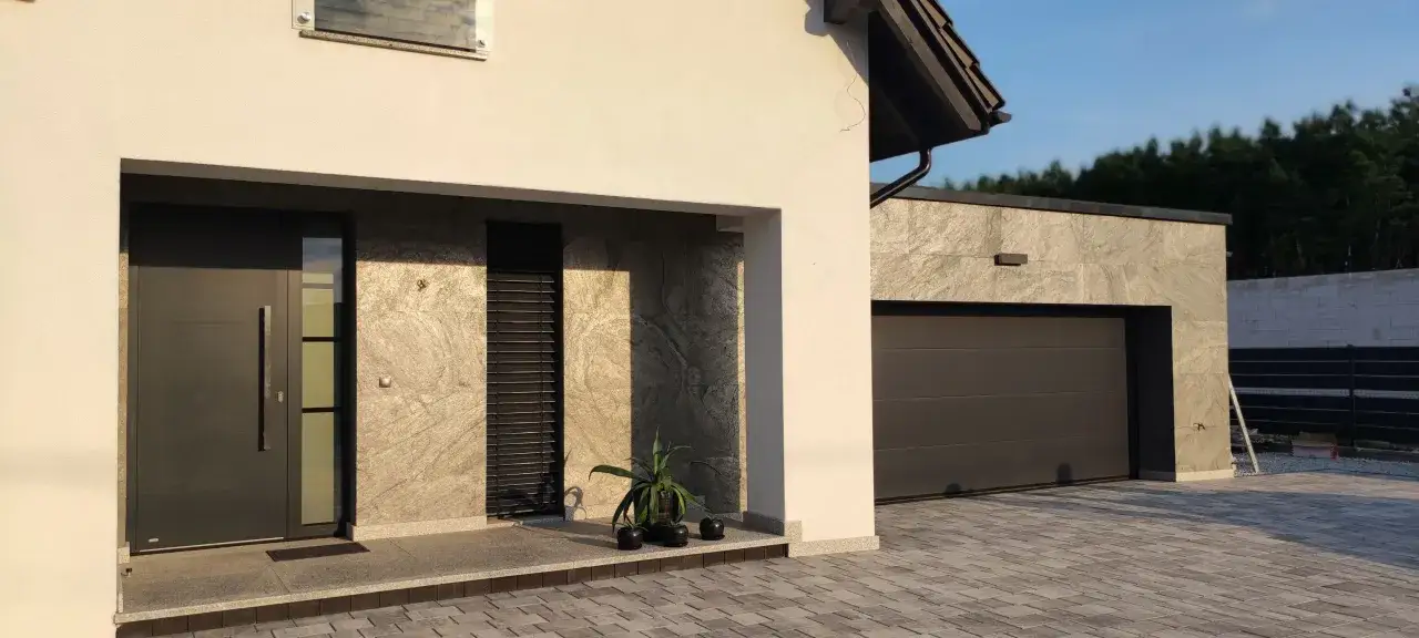 Nowoczesny dom z szarymi płytkami imitującymi kamień, ciemną drzwiami i bramą garażową.