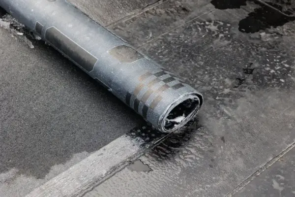 Zrolowana papa na zimno, z charakterystycznym wzorem szachownicy, leży na mokrej, ciemnej powierzchni.